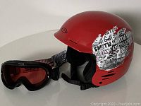 Front view of red Smith helmet with large black and white graphic sticker and Bolle ski goggles with red tinted lenses and black frame next to it.
