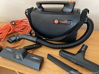 Compact black Hoover PortaPower commercial canister vacuum with shoulder strap, orange extension cord, and four vacuum attachments.