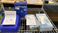 Lot displayed on metal shelf including boxed blood pressure monitor with instruction manual and cuff, non-contact infrared thermometer in plastic bag with manual, and thermometer box showing features.