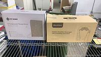 Photo of boxed Fit King air compression therapy device and Maxkare shiatsu shoulder massager box side by side on metal rack.