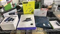 Photo showing all boxed items in the lot on a wire rack including wireless charger, alarm clock, earbuds, keyboard case, audio receiver and headset.