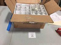 Box containing multiple clear glass votive candle holders with white candles inside, arranged in compartments.