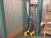 Wide view showing green metal shelving rack holding yellow gas blowers and several long-handled cleaning tools including brooms and mops.