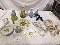 Full view of all Beatrix Potter dishes and figurines displayed on white cloth, showing coffee pot, teacups, plates, figurines including cat figurine that has a cracked base.