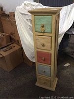 Full view of tall decorative wooden pedestal chest with 5 drawers, each drawer has a colored distressed front and wooden knob.