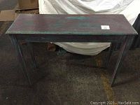 Front view of a wooden console table with multi-color teal and brown distressed paint finish on a concrete floor.