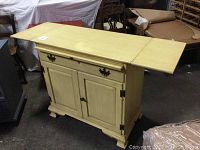 Yellow wooden cabinet with fold-up sides open and slide-out desk surface extended, showing detail of drawer and doors.