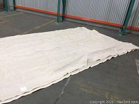Large beige garden patio shade cloth laid out on floor in warehouse setting, showing overall size and condition.