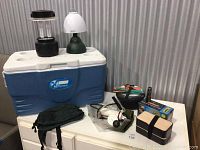 Photo showing blue Xtreme cooler with two camping lanterns on top, black cooking pan, cooking utensils, water filter pump, bento box and radio box on white surface.