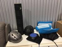 Photo showing two bicycle helmets, black foam roller, blue acupressure mat, blue balance disc, and black neck/back massager pillow placed on a table.