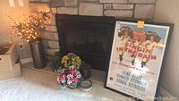 Wide shot showing the framed Singin' in the Rain movie poster leaning against the fireplace alongside artificial floral arrangements and a candle.