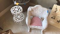 White children's wicker rocking chair with cushion and one white iron plant stand with floral design beside it