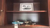 Two metal model cars displayed on a wooden shelf beneath a framed print illustration of Chevrolet cars.