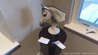 Horse head sculpture on marble base placed on the round top of the plant stand near a window.