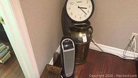 Photo showing the tall brass milk can, atomic clock resting above it, and Oscillarte ceramic heater in corner by wall and baseboard.