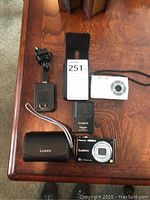 Layout of Lumix and Kodak digital cameras with chargers and accessories on wood surface.