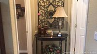 Overview including large ornate metal wall art, small black wooden table, two tall decorative wooden boxes, a small figurine, a candle holder, and a clear glass table lamp with beige shade.
