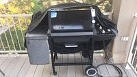 Natural gas Weber grill with cover, shown outdoor on balcony or porch, includes black cover, two wheels, side tray on left side.