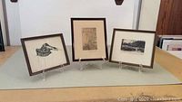 Three framed art pieces arranged side by side on a light surface, showing a pen and ink bird drawing, a print of a road with birch trees by Wallace Nutting, and a framed postcard.