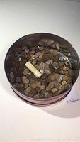 Round container filled with numerous loose copper and steel wheat pennies with at least one rolled paper wrap packet.