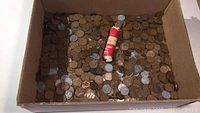 Box filled with an unsorted mix of copper and steel pennies including a red and white wrapped penny roll.