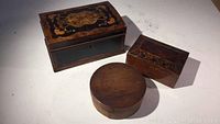 Group view showing all three small wooden boxes: one large decorative burlwood inlay box, one smaller rectangular box with hinged lid, and one round shaker box.