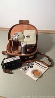 Minolta Zoom 8 movie camera inside brown leather carrying case with user manual, orange filter, and Minolta 16 Model P mini camera with its case in front