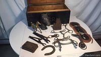Overall view of the collection arranged on a white table showing the open wooden trunk containing multiple equestrian items including bits, brass horse, horseshoe, leather strap, and others.