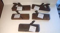 Five antique wood block planes laid out on a white surface showing distinct handles and wooden bodies with labels attached.