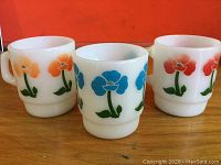 Three vintage FireKing milk glass mugs displayed with orange, blue, and red floral designs on their sides.