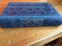 Side view of antique hardcover book showing dark blue cover with embossed pattern and gold embossed title and design on spine.