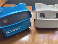 Blue and white vintage View-Master viewers on a wooden surface, showing front side with lenses and brand marking.