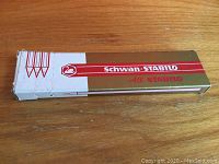Closed gold and white Schwan-Stabilo pencil box with red logo, edges show light wear.