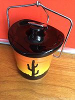 Front view of vintage ceramic sealer jar with black top lid and metal handle against an orange background.