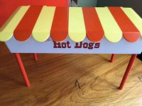 Front angle view of vintage hot dog stand model showing orange and yellow striped canopy and orange legs on wooden surface.