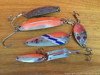 Photo showing six different vintage fishing spoon lures with varying shapes, sizes, paint, and hooks, placed on a wooden surface.