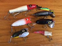 Seven vintage top water plug fishing lures of various sizes, colors, and metal hooks on wooden surface.