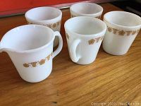 Five white milk glass Pyrex mugs and one creamer with gold floral band near rim on wooden surface.