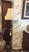 Full view of the tall metal floor lamp with beige fringed lampshade near wallpapered floral wall
