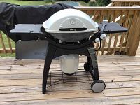 Full view of the Weber gas grill on a wooden deck, showing the white lid, black plastic side tables, legs with wheels, and propane tank holder.