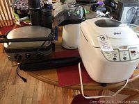 Three small appliances on a dining table including a stainless steel grill, hot air popcorn popper, and bread maker