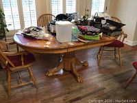 Overview of oval wooden dining table with various household items placed on top. Four spindle back wooden chairs with reddish cushions visible around the table, not included in the lot.
