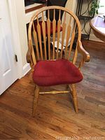 Front view of a wooden Windsor style captain's chair with red cushion, showing vertical slats, armrests, turned legs, and cushion tied with red ribbons.