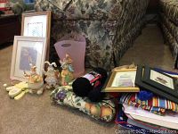 Wide view of the assortment showing framed prints, Easter ceramic figurines, fabric, yarn, and lavender plastic tote on carpeted floor near floral furniture