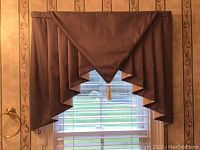 Front view of brown valance with triangular center swag and tassel, hung over window with white blinds and visible wallpaper.