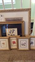 Six framed floral prints stacked and leaning against a wall. Four small gold frames in front showing different flowers. Two larger frames behind with visible matting and floral images.
