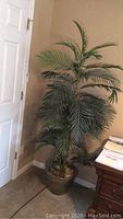 Tall artificial palm plant with multiple layers of plastic green fronds placed inside a brown decorative pot, positioned against a wall beside a door and a wooden furniture item.