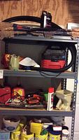 Full shelving unit showing four particle board shelves on a gray metal frame, loaded with various items including a shop vacuum, gas cans, battery charger, and other tools and supplies.