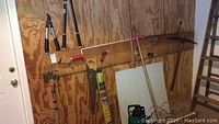 Various garden hand tools including loppers, pruners, sledge hammer, saw hanging on wood wall. Also wooden bungee ladder of approximately 72 inches mounted on wall.
