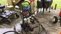 EX-Cell gas powered pressure washer with coiled high-pressure hose and red-handled spray gun. Mounted on black metal frame with wheels. Heavy duty outdoor equipment.
